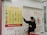 A person wearing a mask stands in front of a currency exchange board displaying various exchange rates between different currencies such as USD, EUR, JPY, and more. The board is yellow with red and black text, accompanied by several national flags. The person appears to be holding a small tray and pointing at the board, likely indicating or discussing the rates. The setting seems to be inside a financial service office, with additional informational posters in the background written in Chinese and English.