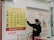 A person wearing a mask stands in front of a currency exchange board displaying various exchange rates between different currencies such as USD, EUR, JPY, and more. The board is yellow with red and black text, accompanied by several national flags. The person appears to be holding a small tray and pointing at the board, likely indicating or discussing the rates. The setting seems to be inside a financial service office, with additional informational posters in the background written in Chinese and English.