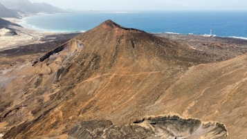A vast, rugged volcanic landscape with a prominent brown and reddish volcano in the foreground. Surrounding the volcano is barren terrain transitioning into a coastal area, with the blue ocean meeting the sandy beach.