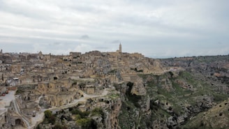 A densely packed ancient city built on a rocky hillside, with numerous stone buildings and narrow streets. The architecture consists of historical structures made from earth-toned materials, blending into the rugged landscape. A prominent tower rises above the cityscape, and the surrounding area is lush with green vegetation.