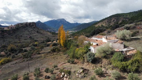 A rustic house with a tiled roof is nestled in a mountainous landscape. The scene includes rolling hills covered in greenery, with a few tall trees showcasing autumn colors. The sky is overcast, casting a soft light over the terrain and enhancing the rugged beauty of the distant mountains.