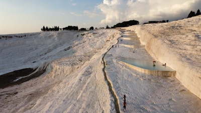 A landscape of white travertine terraces filled with turquoise water, stretching into the distance. People can be seen walking and bathing in the natural pools formed by mineral deposits cascading down the hillside. The sky is partly cloudy with sunlight casting warm hues across the scene.