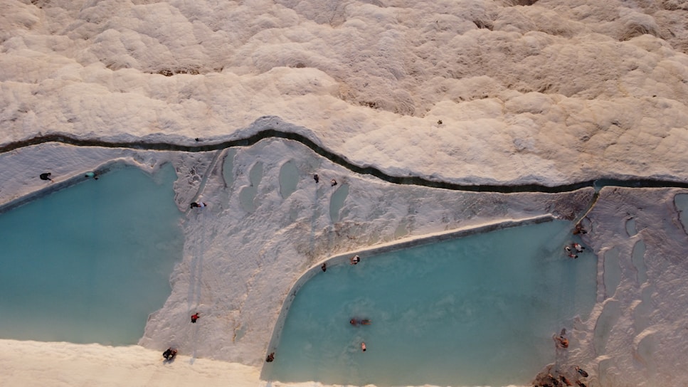 Aerial view of natural mineral terraces filled with turquoise blue water, surrounded by white calcified cliffs. People are seen scattered across the pools, enjoying the serene atmosphere.