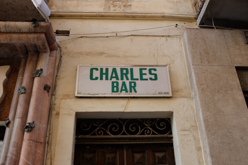A weathered building facade displays a sign reading 'Charles Bar' in green capital letters. The sign is mounted on a light-colored wall, and underneath is a door with decorative ironwork above it. The architecture features stonework with decorative elements.