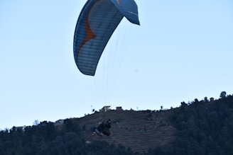 A paraglider is soaring above a hilly landscape, with terraced fields and scattered trees. The scene conveys a sense of adventure and tranquility.