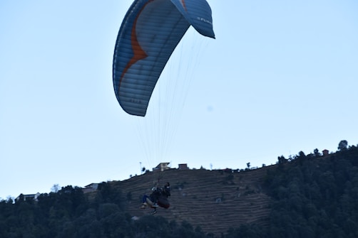 A paraglider is soaring above a hilly landscape, with terraced fields and scattered trees. The scene conveys a sense of adventure and tranquility.