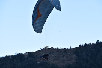 A paraglider is soaring above a hilly landscape, with terraced fields and scattered trees. The scene conveys a sense of adventure and tranquility.