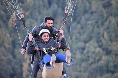 Two people are paragliding, both wearing jackets and safety harnesses. The person in front is wearing a helmet and appears to be smiling, while the person behind is handling the glider. The background consists of blurry, green, forested hills, suggesting they are soaring through a scenic area.