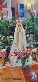 A statue of a woman wearing a white and gold robe with a floral crown, surrounded by a variety of colorful flowers and lush green plants. The background features a religious sign with the word 'Alleluia'.