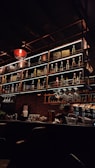 A dimly-lit bar with a warm atmosphere, featuring a well-organized display of various bottles on numerous shelves. The bar counter is adorned with hanging wine glasses, and a bartender can be spotted attending to tasks behind the bar.