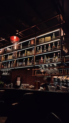A dimly-lit bar with a warm atmosphere, featuring a well-organized display of various bottles on numerous shelves. The bar counter is adorned with hanging wine glasses, and a bartender can be spotted attending to tasks behind the bar.