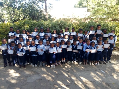 A large group of young children are standing together outdoors, each holding a certificate. The children are dressed in school uniforms consisting of blue and white shirts, vests, and skirts or pants. They are standing in rows, with greenery in the background, suggesting a garden or outdoor area.