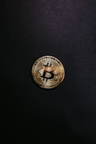 A single gold-colored Bitcoin physical coin is centered against a dark black background. The coin displays a stylized 'B' symbol and circuit-like patterns that are characteristic of cryptocurrency designs.