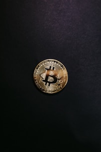 A single gold-colored Bitcoin physical coin is centered against a dark black background. The coin displays a stylized 'B' symbol and circuit-like patterns that are characteristic of cryptocurrency designs.