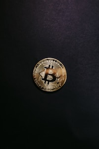A single gold-colored Bitcoin physical coin is centered against a dark black background. The coin displays a stylized 'B' symbol and circuit-like patterns that are characteristic of cryptocurrency designs.