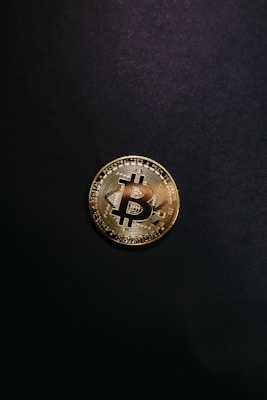 A single gold-colored Bitcoin physical coin is centered against a dark black background. The coin displays a stylized 'B' symbol and circuit-like patterns that are characteristic of cryptocurrency designs.