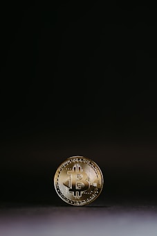 A gold Bitcoin coin is shown standing upright against a minimalistic dark background. The coin features the Bitcoin emblem prominently in the center, surrounded by circuit-like patterns. The lighting creates a subtle glow around the edges, emphasizing the coin&rsquo;s metallic sheen.
