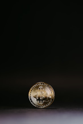 A gold Bitcoin coin is shown standing upright against a minimalistic dark background. The coin features the Bitcoin emblem prominently in the center, surrounded by circuit-like patterns. The lighting creates a subtle glow around the edges, emphasizing the coin’s metallic sheen.