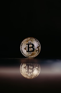 A single Bitcoin coin stands upright on a reflective surface, casting a clear reflection beneath it. The background is a dark black, enhancing the visibility of the coin and its intricate details.
