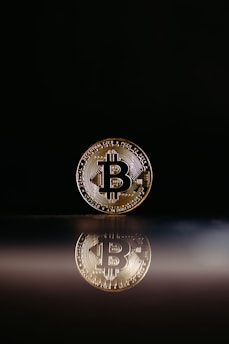 A single Bitcoin coin stands upright on a reflective surface, casting a clear reflection beneath it. The background is a dark black, enhancing the visibility of the coin and its intricate details.