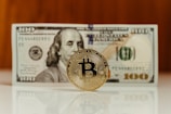 A Bitcoin coin stands upright on a reflective surface in front of a hundred-dollar bill, featuring the image of Benjamin Franklin. The focus is on the Bitcoin, with the bill slightly out of focus in the background.