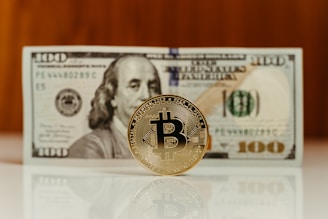 A Bitcoin coin stands upright on a reflective surface in front of a hundred-dollar bill, featuring the image of Benjamin Franklin. The focus is on the Bitcoin, with the bill slightly out of focus in the background.