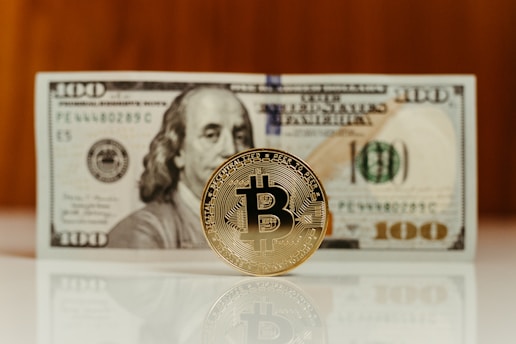 A Bitcoin coin stands upright on a reflective surface in front of a hundred-dollar bill, featuring the image of Benjamin Franklin. The focus is on the Bitcoin, with the bill slightly out of focus in the background.