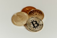 A close-up image of physical representations of cryptocurrencies, including Bitcoin, Ethereum, and Cardano coins, arranged on a plain light-colored surface.