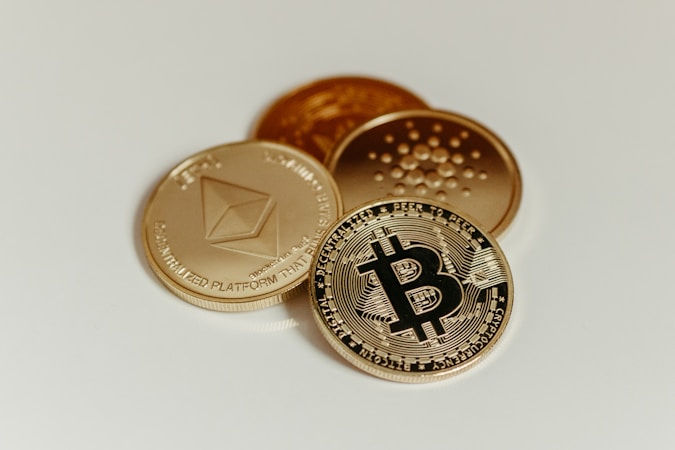 A close-up image of physical representations of cryptocurrencies, including Bitcoin, Ethereum, and Cardano coins, arranged on a plain light-colored surface.