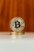 A shiny golden Bitcoin coin stands vertically on top of a small stack of similar coins. The coin's intricate details and 'B' symbol are clearly visible. The background is blurred with a warm, wooden tone, and the reflection of the coins is visible on the glossy surface below.