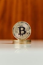A shiny golden Bitcoin coin stands vertically on top of a small stack of similar coins. The coin's intricate details and 'B' symbol are clearly visible. The background is blurred with a warm, wooden tone, and the reflection of the coins is visible on the glossy surface below.