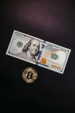 A hundred dollar bill featuring Benjamin Franklin is displayed against a dark background. Below and slightly to the left, a physical representation of a Bitcoin coin is positioned. The Bitcoin has a golden color with a prominent Bitcoin logo in the center.