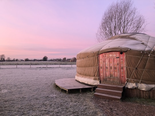A traditional yurt with ornate red and pink wooden door at the edge of a frost-covered field. The sky is tinged with pink hues, suggesting early morning or late evening. Bare trees and distant buildings are visible on the horizon.