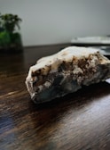 A close-up photo of a rough mineral or crystal rock placed on a dark wooden surface. The rock has various textures and colors, including white, brown, and gray areas. In the background, there is a blurry view of a plant and possibly a book or a stack of papers.