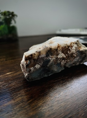 A close-up photo of a rough mineral or crystal rock placed on a dark wooden surface. The rock has various textures and colors, including white, brown, and gray areas. In the background, there is a blurry view of a plant and possibly a book or a stack of papers.