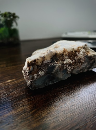 A close-up photo of a rough mineral or crystal rock placed on a dark wooden surface. The rock has various textures and colors, including white, brown, and gray areas. In the background, there is a blurry view of a plant and possibly a book or a stack of papers.