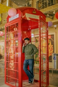 A man leans casually against the wall of a traditional red British telephone booth while talking on a mobile phone. The booth is situated indoors, surrounded by decorative hanging lanterns. He is wearing a green jacket and jeans. The background features an industrial-style interior with large paintings and a stainless steel trash bin.