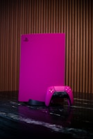 A gaming console and controller, both in a vibrant pink color, are placed on a dark reflective surface. The background consists of vertical wooden panels, providing a warm contrast to the bright gaming equipment.