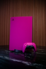A gaming console and controller, both in a vibrant pink color, are placed on a dark reflective surface. The background consists of vertical wooden panels, providing a warm contrast to the bright gaming equipment.