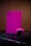 A gaming console and controller, both in a vibrant pink color, are placed on a dark reflective surface. The background consists of vertical wooden panels, providing a warm contrast to the bright gaming equipment.