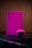 A gaming console and controller, both in a vibrant pink color, are placed on a dark reflective surface. The background consists of vertical wooden panels, providing a warm contrast to the bright gaming equipment.