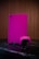 A gaming console and controller, both in a vibrant pink color, are placed on a dark reflective surface. The background consists of vertical wooden panels, providing a warm contrast to the bright gaming equipment.
