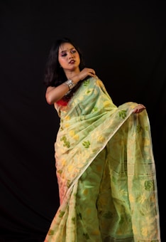A person wearing a light-colored saree with floral patterns stands against a dark background. The individual has long black hair and is adorned with traditional jewelry, including bangles and earrings.