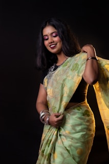 A smiling person wearing a light green saree with floral patterns, accessorized with bangles and a black blouse. The individual has long black hair and is standing against a dark background.
