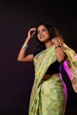 A woman wearing a green saree and black blouse stands against a dark background. She is adorned with bangles and her hair is styled naturally, with a smile on her face and one hand raised near her head.