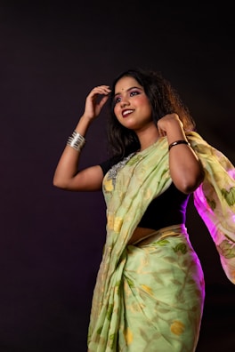 A woman wearing a green saree and black blouse stands against a dark background. She is adorned with bangles and her hair is styled naturally, with a smile on her face and one hand raised near her head.