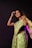 A woman wearing a green saree and black blouse stands against a dark background. She is adorned with bangles and her hair is styled naturally, with a smile on her face and one hand raised near her head.