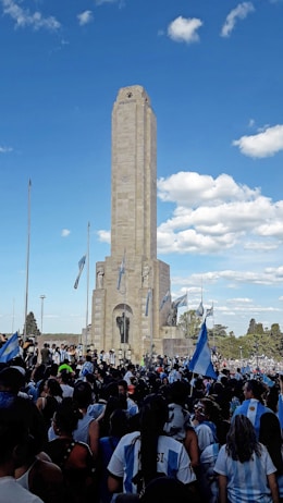 A large crowd gathers in front of a tall monument adorned with Argentine flags and statues. The people, many wearing blue and white clothing, create a lively atmosphere under a clear blue sky with scattered clouds. Trees and additional flags surround the structure.