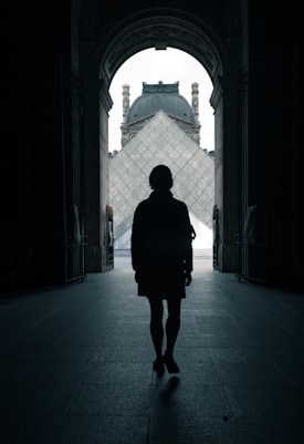 A silhouette of a person standing in an archway, facing the iconic glass pyramid of the Louvre Museum with its historical architecture visible in the background.
