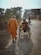 An older adult dressed in orange walks beside a person in a wheelchair on a rural road. The road is lined with trees and buildings. Other people are seen riding motorcycles further ahead.